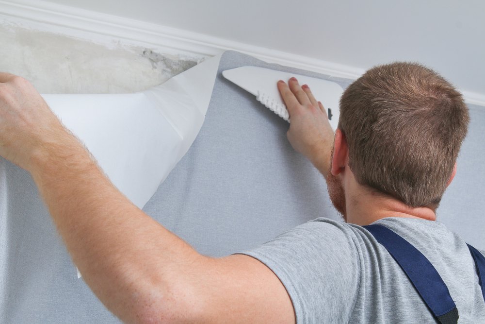 Een rol renovlies wordt aangebracht op een muur door een persoon met behulp van een behangborstel en andere benodigde gereedschappen.