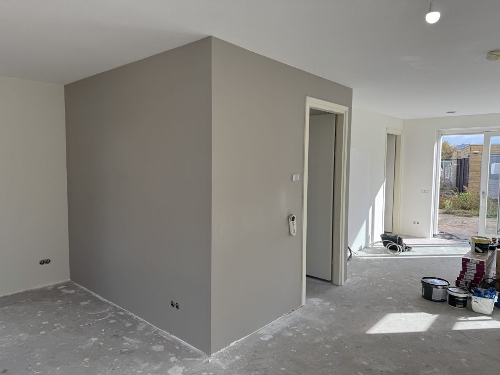 Strak afgewerkte grijze wand met scherpe hoeken in een nieuwbouwwoning, verfemmers op de vloer