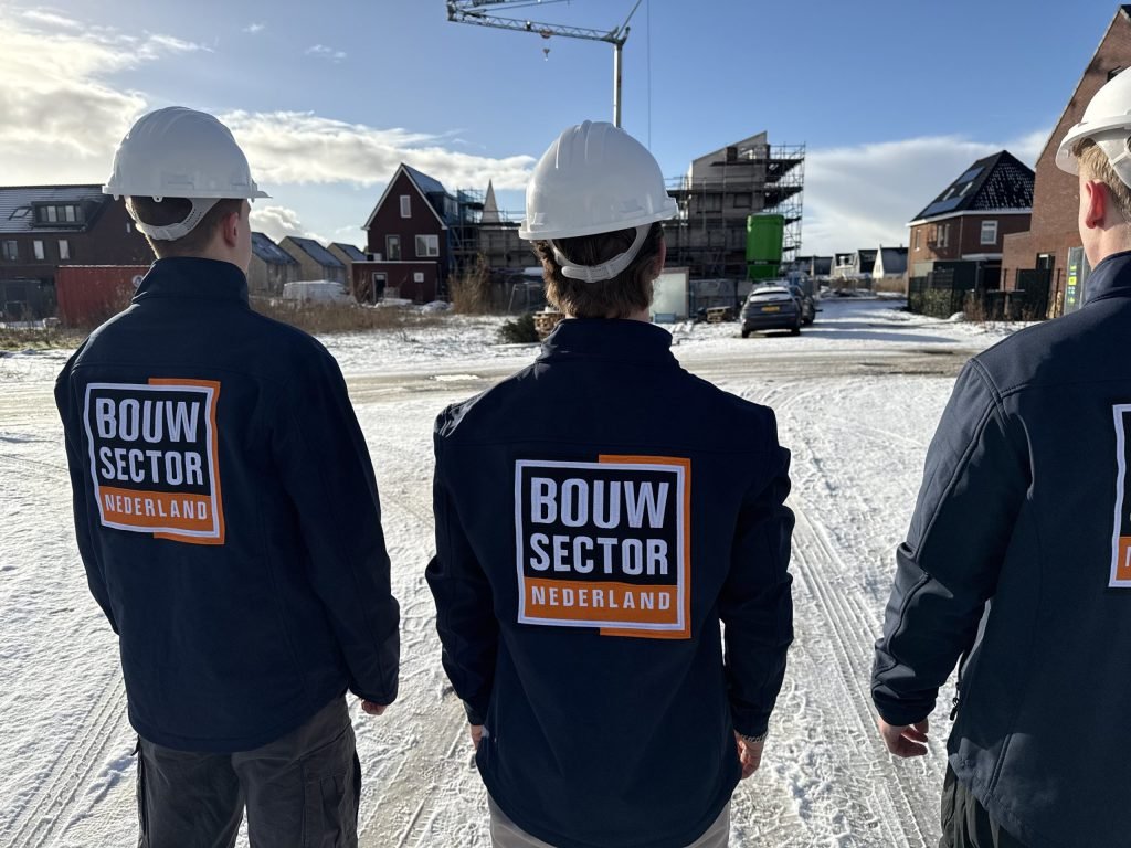 Drie vakmensen in werkkleding met het logo van Bouwsector Nederland op de rug voor een rij huizen in aanbouw